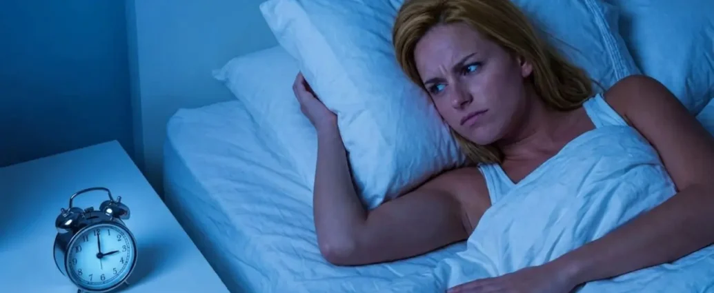 Una donna bionda sveglia a letto nel cuore della notte con espressione preoccupata guarda la sveglia che segna le 3:00. Scena illuminata da luce fredda bluastra.