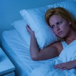 Una donna bionda sveglia a letto nel cuore della notte con espressione preoccupata guarda la sveglia che segna le 3:00. Scena illuminata da luce fredda bluastra.
