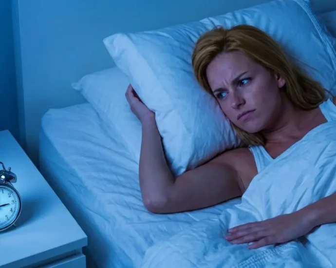Una donna bionda sveglia a letto nel cuore della notte con espressione preoccupata guarda la sveglia che segna le 3:00. Scena illuminata da luce fredda bluastra.