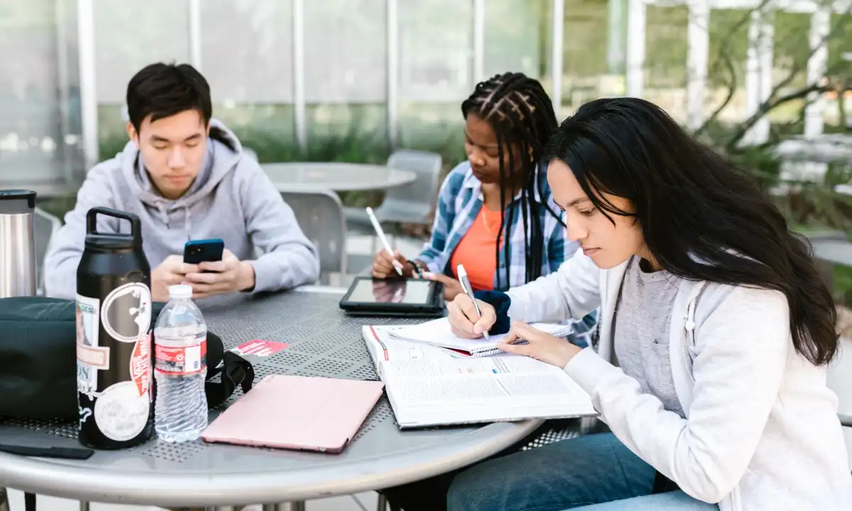 Un gruppo di studenti seduti a un tavolo all'aperto mentre studiano, scrivono appunti e consultano libri e dispositivi digitali.